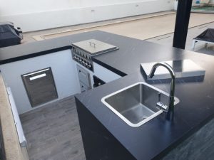 מטבח חוץ משולב בר ישיבה שיש למינם פינתי. מאובזר בגריל גז, כיור, כיריים גז וארונות אחסון